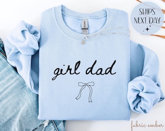 May include: A light blue sweatshirt with the words "girl dad" in black script and a bow graphic. A silver necklace with a small pendant is on the sweatshirt. The sweatshirt is folded on top of a pair of blue jeans.