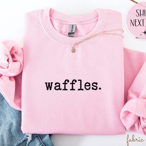 K&ouml;nnte beinhalten: Ein hellrosa Sweatshirt mit dem Wort "waffles." in schwarzer Schrift. Das Sweatshirt ist gefaltet, wobei die &Auml;rmel sichtbar sind. Eine goldene Halskette mit einem kleinen Anh&auml;nger ist dar&uuml;ber drapiert. Die Worte "SHIPS NEXT DAY" stehen in einem rosa Kreis.