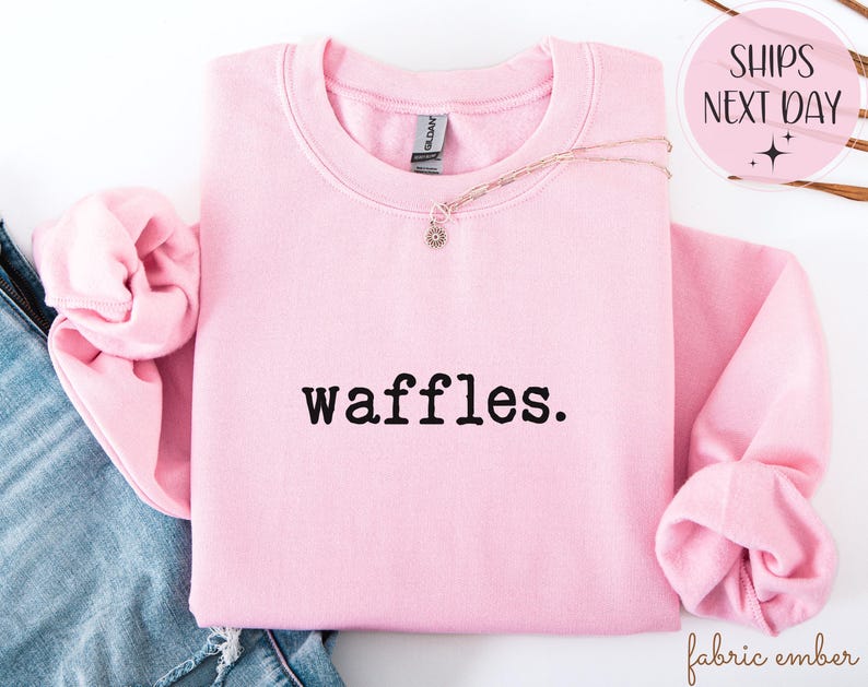 K&ouml;nnte beinhalten: Ein hellrosa Sweatshirt mit dem Wort "waffles." in schwarzer Schrift. Das Sweatshirt ist gefaltet, wobei die &Auml;rmel sichtbar sind. Eine goldene Halskette mit einem kleinen Anh&auml;nger ist dar&uuml;ber drapiert. Die Worte "SHIPS NEXT DAY" stehen in einem rosa Kreis.