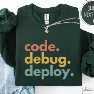 May include: A dark green sweatshirt with the words "code. debug. deploy." in orange, yellow, and teal. A silver necklace with a pendant is draped across the sweatshirt. The sweatshirt is folded on top of a pair of blue jeans.