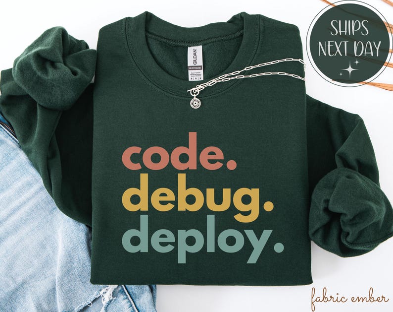 May include: A dark green sweatshirt with the words "code. debug. deploy." in orange, yellow, and teal. A silver necklace with a pendant is draped across the sweatshirt. The sweatshirt is folded on top of a pair of blue jeans.