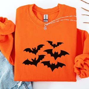 Puede incluir: Sudadera naranja con cuello redondo y mangas largas. La parte delantera de la camisa presenta un gr&aacute;fico de murci&eacute;lagos negros. Un collar plateado con un colgante circular est&aacute; sobre la camisa.