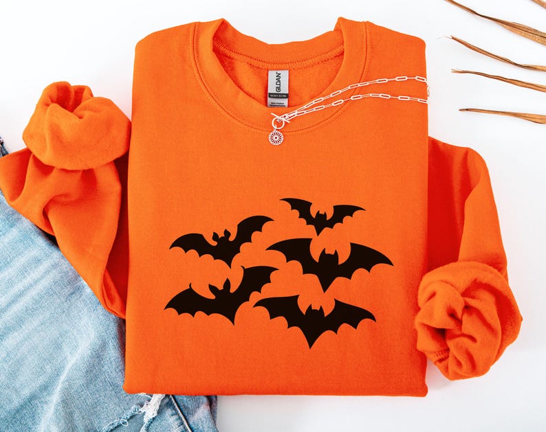 Puede incluir: Sudadera naranja con cuello redondo y mangas largas. La parte delantera de la camisa presenta un gr&aacute;fico de murci&eacute;lagos negros. Un collar plateado con un colgante circular est&aacute; sobre la camisa.