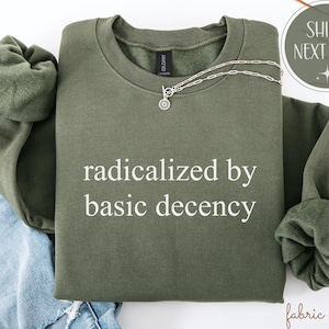 Op de afbeelding: Een olijfgroene sweatshirt met de tekst "radicalized by basic decency" in het wit. Een zilveren ketting met een cirkelvormige hanger is over de voorkant gedrapeerd. De sweatshirt is op blauwe denim gevouwen.