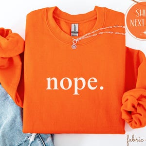 May include: Bright orange sweatshirt with the word "nope." in white block letters. The sweatshirt has a crew neck and long sleeves, with the sleeves rolled up. A silver necklace with a sun pendant is draped across the front.