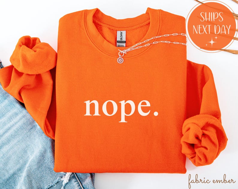 May include: Bright orange sweatshirt with the word "nope." in white block letters. The sweatshirt has a crew neck and long sleeves, with the sleeves rolled up. A silver necklace with a sun pendant is draped across the front.