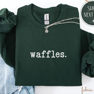 K&ouml;nnte beinhalten: Ein dunkelgr&uuml;nes Sweatshirt mit dem wei&szlig;en Aufdruck "waffles". Eine silberne Halskette mit einem kreisf&ouml;rmigen Anh&auml;nger liegt &uuml;ber der Vorderseite. Das Sweatshirt ist auf einer Jeans gefaltet.