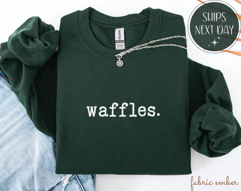 K&ouml;nnte beinhalten: Ein dunkelgr&uuml;nes Sweatshirt mit dem wei&szlig;en Aufdruck "waffles". Eine silberne Halskette mit einem kreisf&ouml;rmigen Anh&auml;nger liegt &uuml;ber der Vorderseite. Das Sweatshirt ist auf einer Jeans gefaltet.
