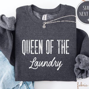 May include: Dark gray sweatshirt with the text "QUEEN OF THE LAUNDRY" in white. A silver necklace with a pendant is draped across the front. The sweatshirt is folded on top of a pair of blue jeans.