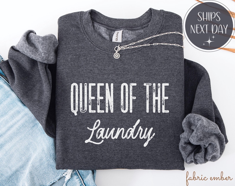 May include: Dark gray sweatshirt with the text "QUEEN OF THE LAUNDRY" in white. A silver necklace with a pendant is draped across the front. The sweatshirt is folded on top of a pair of blue jeans.