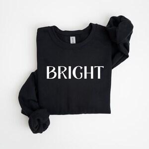 May include: Black crew neck sweatshirt with the word "BRIGHT" printed in large, white capital letters across the front. The sweatshirt is folded neatly on a white surface. The sleeves are folded in.