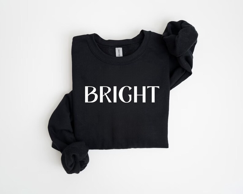 May include: Black crew neck sweatshirt with the word "BRIGHT" printed in large, white capital letters across the front. The sweatshirt is folded neatly on a white surface. The sleeves are folded in.