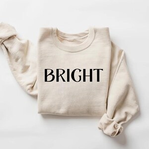 May include: A cream-coloured sweatshirt with the word "BRIGHT" printed in black across the front. The sleeves are folded at the wrists. The sweatshirt is made of a soft material and is folded neatly.
