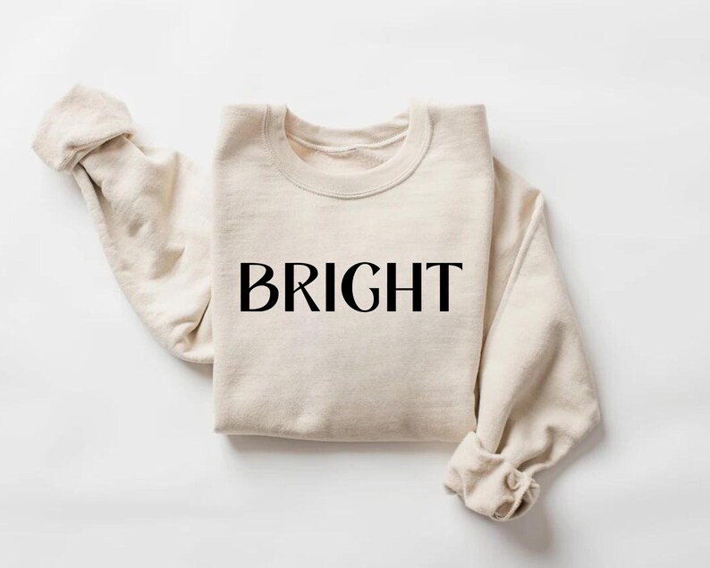 May include: A cream-coloured sweatshirt with the word "BRIGHT" printed in black across the front. The sleeves are folded at the wrists. The sweatshirt is made of a soft material and is folded neatly.