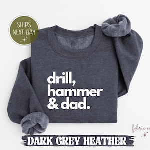 Könnte beinhalten: Dunkelgraues Heather-Sweatshirt mit dem weißen Text "drill, hammer & dad". Das Sweatshirt hat einen Rundhalsausschnitt und lange Ärmel. Die Worte "Dark Grey Heather" sind am unteren Rand des Bildes aufgedruckt.
