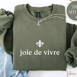 May include: An olive green sweatshirt with the phrase "joie de vivre" in white, and a fleur-de-lis symbol. A silver necklace with a circular pendant rests on the sweatshirt. The sweatshirt has a crew neck and long sleeves.