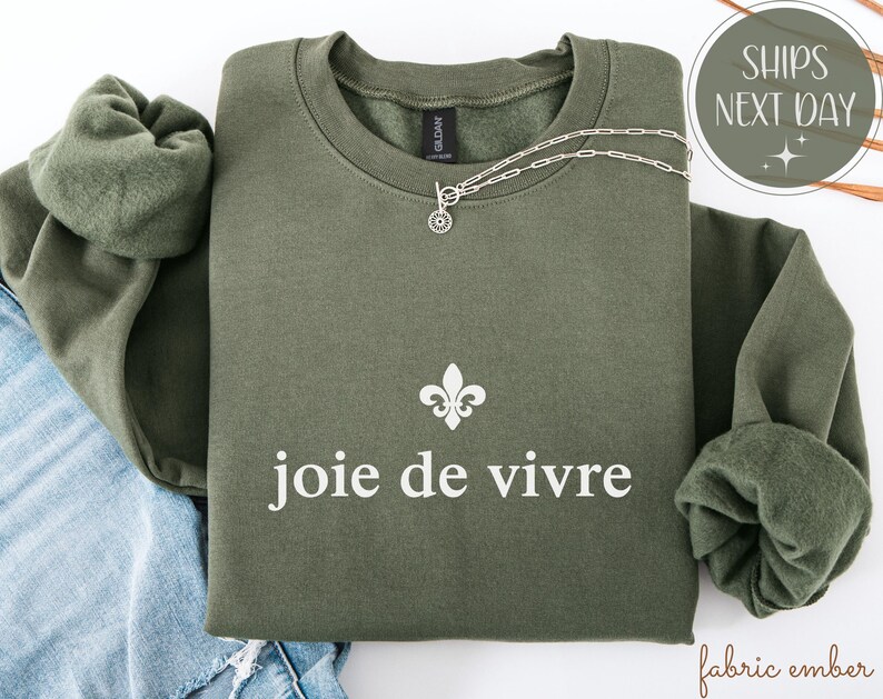 May include: An olive green sweatshirt with the phrase "joie de vivre" in white, and a fleur-de-lis symbol. A silver necklace with a circular pendant rests on the sweatshirt. The sweatshirt has a crew neck and long sleeves.