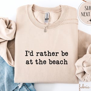 May include: A beige sweatshirt with the text "I'd rather be at the beach" in black. A silver necklace with a pendant is on the sweatshirt. The sweatshirt is folded with a pair of blue jeans and a rolled-up sleeve.