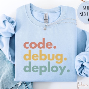 May include: A pale blue sweatshirt featuring the words "code. debug. deploy." in orange, yellow, and green. A silver necklace with a small pendant rests on the sweatshirt. The sweatshirt is folded on top of blue denim jeans.