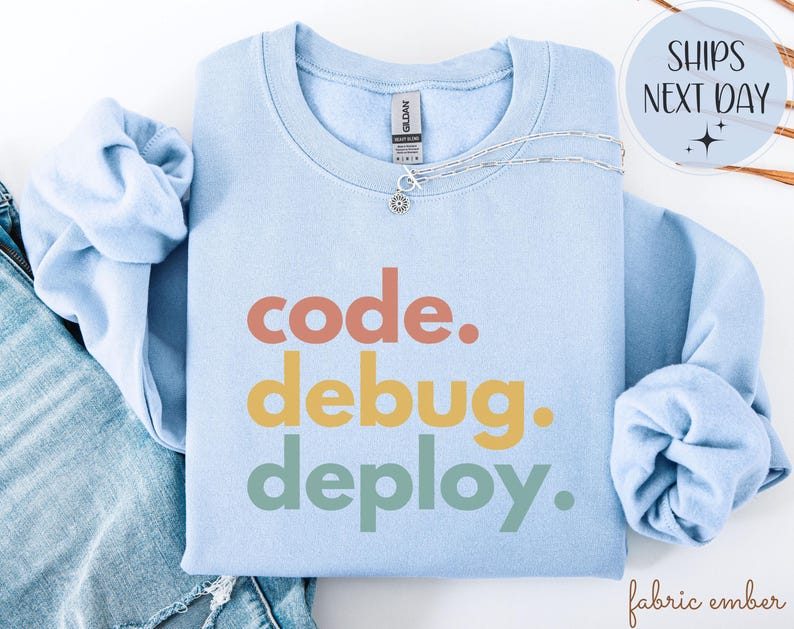 May include: A pale blue sweatshirt featuring the words "code. debug. deploy." in orange, yellow, and green. A silver necklace with a small pendant rests on the sweatshirt. The sweatshirt is folded on top of blue denim jeans.