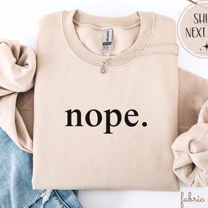 May include: A beige sweatshirt with the word "nope." printed in black. The sweatshirt is folded, with the sleeves and collar visible. A silver necklace with a small pendant rests on the fabric.
