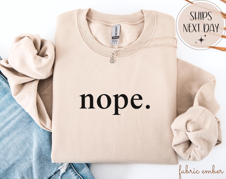 May include: A beige sweatshirt with the word "nope." printed in black. The sweatshirt is folded, with the sleeves and collar visible. A silver necklace with a small pendant rests on the fabric.