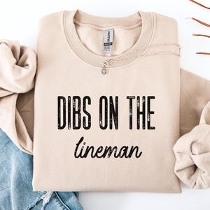 Op de afbeelding: Een beige sweatshirt met de tekst "DIBS ON THE lineman" in het zwart. De sweatshirt is opgevouwen met een spijkerbroek en een ketting. De sweatshirt is gemaakt van een zacht materiaal.