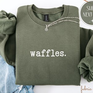 K&ouml;nnte beinhalten: Ein olivgr&uuml;nes Sweatshirt mit dem wei&szlig;en Aufdruck "waffles". Eine silberne Halskette mit einem kreisf&ouml;rmigen Anh&auml;nger ist &uuml;ber die Vorderseite drapiert. Das Sweatshirt ist &uuml;ber einer Jeans gefaltet.