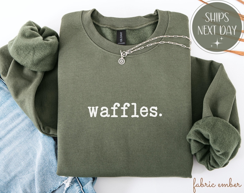 K&ouml;nnte beinhalten: Ein olivgr&uuml;nes Sweatshirt mit dem wei&szlig;en Aufdruck "waffles". Eine silberne Halskette mit einem kreisf&ouml;rmigen Anh&auml;nger ist &uuml;ber die Vorderseite drapiert. Das Sweatshirt ist &uuml;ber einer Jeans gefaltet.