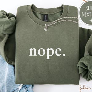May include: An olive green sweatshirt with the word "nope" printed in white. A silver necklace with a circular pendant rests on the sweatshirt. The sleeves are folded, and the sweatshirt is on top of blue denim.