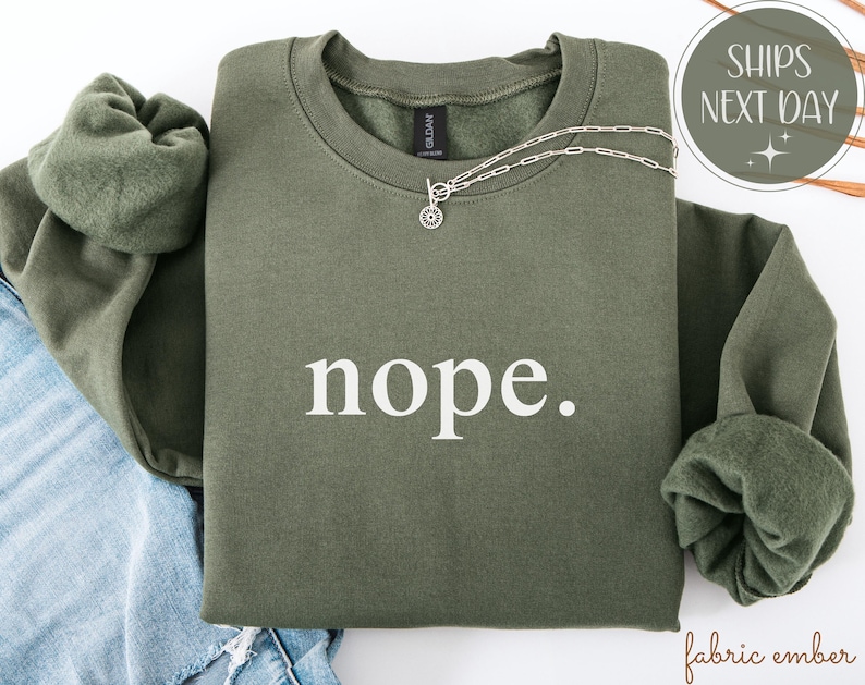 May include: An olive green sweatshirt with the word "nope" printed in white. A silver necklace with a circular pendant rests on the sweatshirt. The sleeves are folded, and the sweatshirt is on top of blue denim.