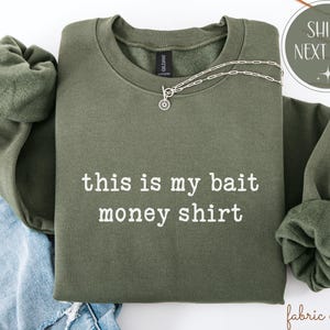 May include: An olive green sweatshirt with the text "this is my bait money shirt" in white. A silver necklace with a circular pendant is draped across the shirt. The sweatshirt is folded on top of a pair of blue jeans.