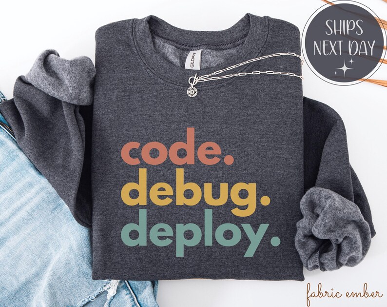 May include: A charcoal grey sweatshirt featuring the text "code. debug. deploy." in orange, yellow, and teal. A silver necklace with a pendant is draped across the shirt. The image also includes a pair of blue jeans.