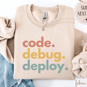 May include: A beige sweatshirt featuring the words "code. debug. deploy." in coral, yellow, and teal. The sweatshirt has a crew neck and long sleeves. A silver necklace with a small pendant is draped on the sweatshirt.