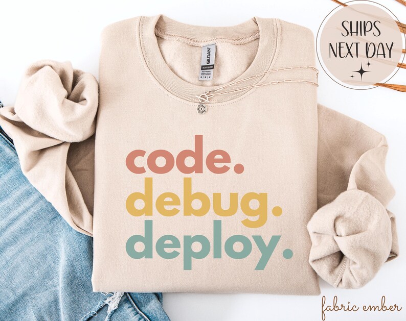 May include: A beige sweatshirt featuring the words "code. debug. deploy." in coral, yellow, and teal. The sweatshirt has a crew neck and long sleeves. A silver necklace with a small pendant is draped on the sweatshirt.