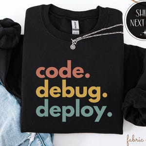 Puede incluir: Sudadera negra con las palabras "code. debug. deploy." en un esquema de colores retro. Un collar plateado con un pequeño colgante está drapeado sobre la sudadera. La sudadera está doblada sobre unos vaqueros azules.