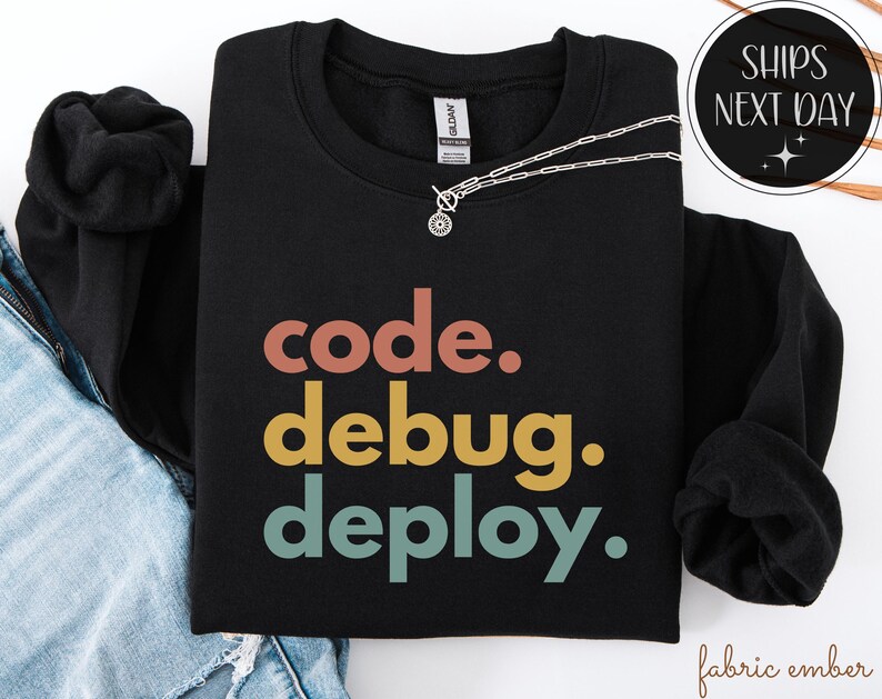 May include: Black sweatshirt featuring the words "code. debug. deploy." in a retro colour scheme. A silver necklace with a small pendant is draped over the sweatshirt. The sweatshirt is folded on top of a pair of blue jeans.