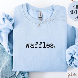 K&ouml;nnte beinhalten: Hellblaues Sweatshirt mit dem Wort "waffles." in Schwarz aufgedruckt. Eine silberne Halskette mit einem kleinen Anh&auml;nger befindet sich auf dem Sweatshirt. Das Sweatshirt ist gefaltet, die &Auml;rmel sind sichtbar. Die Worte "Ships Next Day" stehen in einem Kreis.