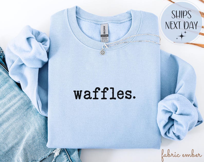 K&ouml;nnte beinhalten: Hellblaues Sweatshirt mit dem Wort "waffles." in Schwarz aufgedruckt. Eine silberne Halskette mit einem kleinen Anh&auml;nger befindet sich auf dem Sweatshirt. Das Sweatshirt ist gefaltet, die &Auml;rmel sind sichtbar. Die Worte "Ships Next Day" stehen in einem Kreis.