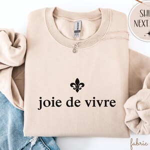 May include: A beige sweatshirt featuring the phrase "joie de vivre" in black, with a fleur-de-lis symbol. A delicate necklace with a pendant is draped on the sweatshirt. The sweatshirt is folded with the sleeves tucked in.