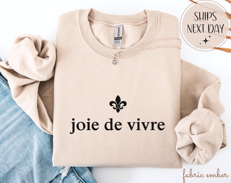 May include: A beige sweatshirt featuring the phrase "joie de vivre" in black, with a fleur-de-lis symbol. A delicate necklace with a pendant is draped on the sweatshirt. The sweatshirt is folded with the sleeves tucked in.
