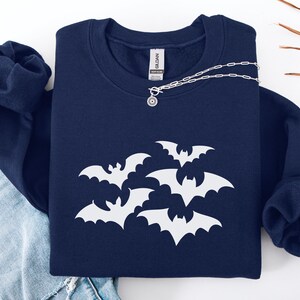 Puede incluir: Una sudadera azul marino con un dise&ntilde;o de murci&eacute;lagos blancos. Un collar plateado con un colgante circular est&aacute; sobre la sudadera. La sudadera est&aacute; doblada sobre un par de vaqueros azules.