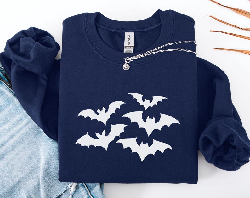 Puede incluir: Una sudadera azul marino con un dise&ntilde;o de murci&eacute;lagos blancos. Un collar plateado con un colgante circular est&aacute; sobre la sudadera. La sudadera est&aacute; doblada sobre un par de vaqueros azules.
