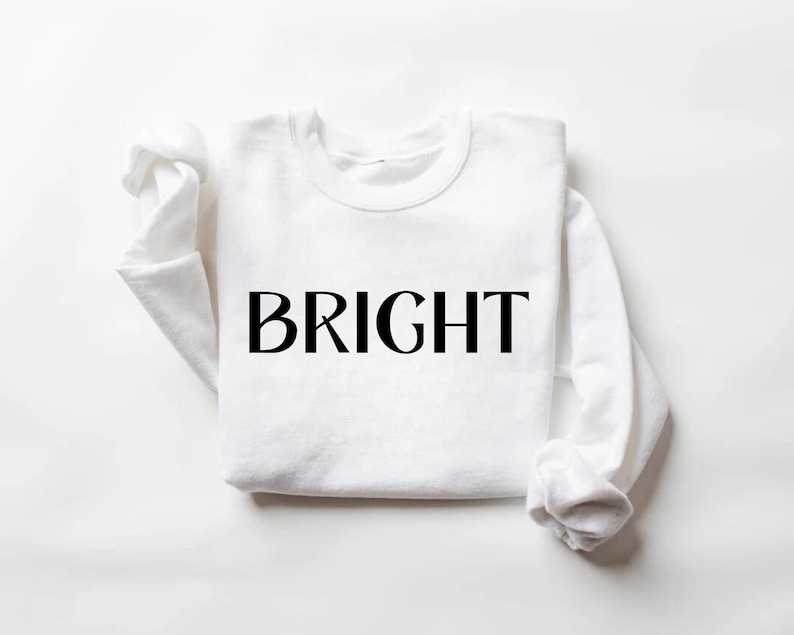 May include: A white crew neck sweatshirt with the word "BRIGHT" printed in large, black capital letters across the front. The sleeves are folded up. The sweatshirt is made of a soft material.