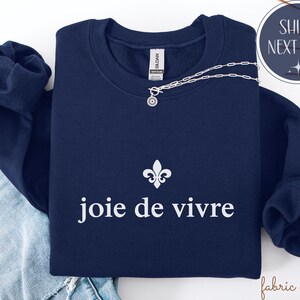 May include: A navy blue sweatshirt featuring the words "joie de vivre" in white, accompanied by a fleur-de-lis symbol. A silver necklace with a circular pendant is placed on the sweatshirt. The sweatshirt is folded on blue jeans.