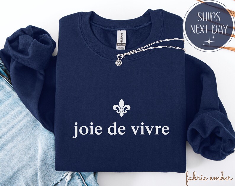 May include: A navy blue sweatshirt featuring the words "joie de vivre" in white, accompanied by a fleur-de-lis symbol. A silver necklace with a circular pendant is placed on the sweatshirt. The sweatshirt is folded on blue jeans.