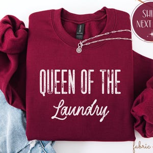 May include: A burgundy crewneck sweatshirt with the text "QUEEN OF THE LAUNDRY" in white. A silver necklace with a circular pendant is draped across the sweatshirt. The sweatshirt is folded on top of blue denim.