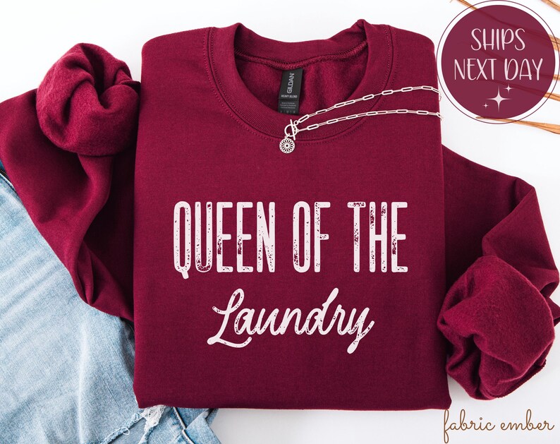 May include: A burgundy crewneck sweatshirt with the text "QUEEN OF THE LAUNDRY" in white. A silver necklace with a circular pendant is draped across the sweatshirt. The sweatshirt is folded on top of blue denim.
