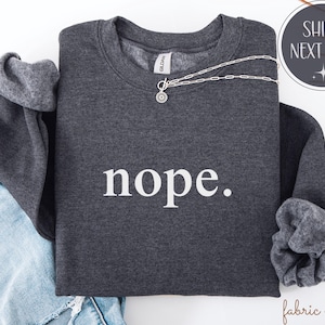 May include: A dark gray sweatshirt with the word "nope" printed in white. A silver necklace with a circular pendant rests on the sweatshirt. The sleeves are folded, revealing a lighter gray lining.