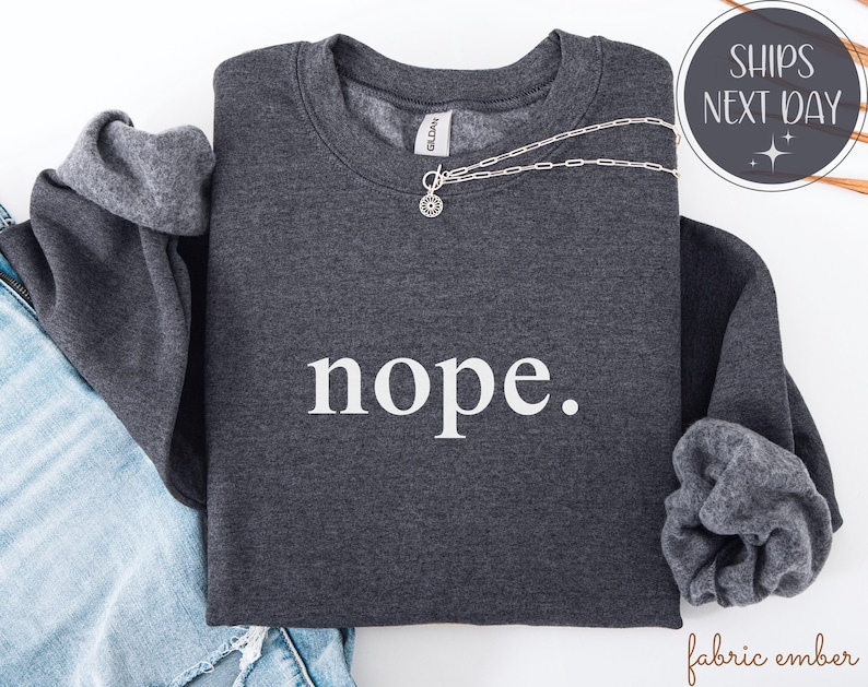 May include: A dark gray sweatshirt with the word "nope" printed in white. A silver necklace with a circular pendant rests on the sweatshirt. The sleeves are folded, revealing a lighter gray lining.